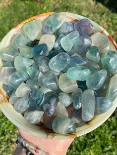 a person holding a bowl full of green and blue rocks in their hand with grass behind it