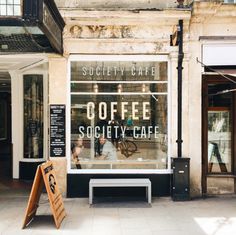 an empty sidewalk in front of a coffee shop with signage on the window and people sitting outside