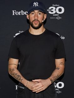 a man with tattoos standing in front of a black backdrop and wearing a baseball cap