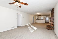 an empty living room with a ceiling fan and brick fireplace in the corner, next to a kitchen