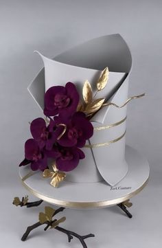 a white cake with purple flowers and gold leaves on it's stand, against a gray background