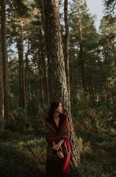 a woman standing next to a tall tree in the middle of a forest wearing a blanket