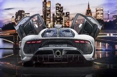 the rear end of a silver sports car with city lights in the background