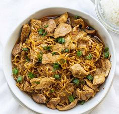 a white bowl filled with noodles and meat on top of a table next to rice