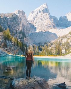 a woman standing on top of a rock next to a lake with mountains in the background