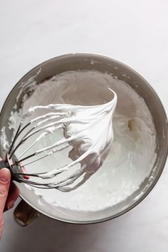 a hand holding a whisk in a metal bowl with white icing on it