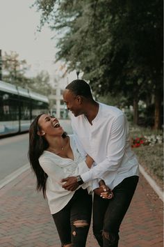 a man and woman laughing while standing next to each other on a brick sidewalk with a train in the background