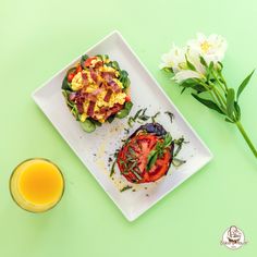 two small sandwiches on a white plate next to a glass of orange juice and some flowers