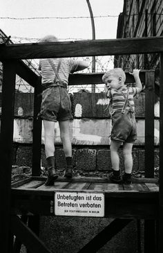 two small children standing on top of a wooden platform in front of a building with barbed wire