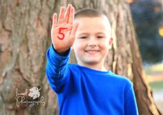 a young boy holding his hand up in the air while standing next to a tree