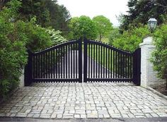 an iron gate and brick walkway leading to a driveway surrounded by greenery on either side