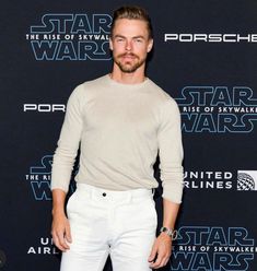 a man standing in front of a star wars logo on a black background wearing white pants and a sweater