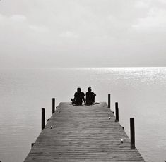 two people sitting on the end of a pier looking out at the water in black and white