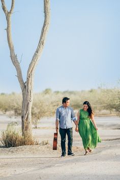 a man and woman holding hands walking down a dirt road with a tree in the background