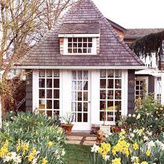 a small house with lots of windows and flowers in the front yard, surrounded by greenery