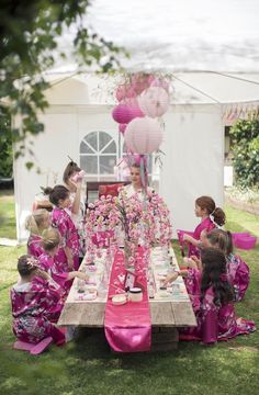 a group of children sitting around a table with pink decorations on it and balloons in the background