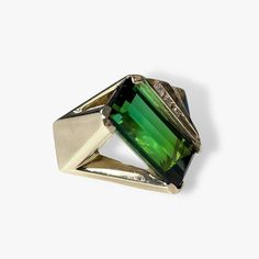 A one-of-a-kind vintage cocktail ring boasting a vibrant elongated emerald cut green tourmaline weighing 16.77 carats set on an angle while accented by 5 round diamonds weighing 0.32 carats set in solid 14k yellow gold with tiered detailing. Complimentary resizing is available up to 3 sizes larger or smaller than the stated size. WEB ID: R603