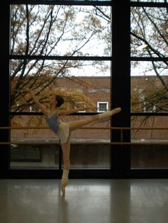 a young ballerina is practicing her moves in front of large windows with trees outside