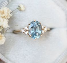 an oval blue topaz and diamond ring sits on a white surface next to flowers