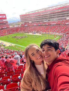 a man and woman taking a selfie in front of a red stadium full of people