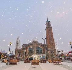 a clock tower is in the middle of a snowy day with benches and lights around it