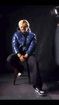 a man is sitting on a chair in front of a black background, wearing a blue jacket and white turban