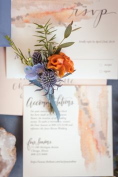 an orange and blue flower is on top of the wedding stationery with greenery