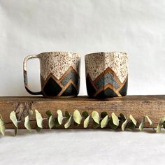 two coffee mugs sitting next to each other on a piece of wood with leaves