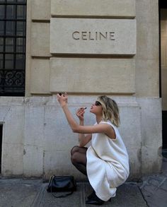 a woman sitting on the ground in front of a building with her hand up to her mouth