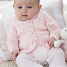 a baby sitting on the floor with a stuffed animal in its lap and wearing a pink sweater