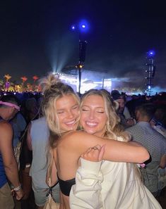 two women hugging each other in front of a crowd at a music festival with bright lights behind them