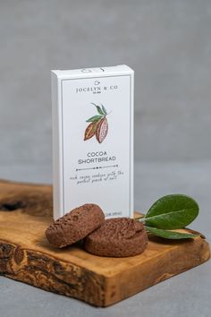 a box of cocoa shortbreads next to two cookies on a wooden board with leaves