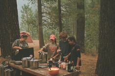 three men standing around a picnic table in the woods with food and drinks on it