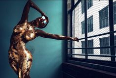 a woman standing in front of a window with her arms outstretched and body painted gold