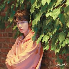 a young man standing next to a brick wall covered in green leaves and wearing an orange hoodie