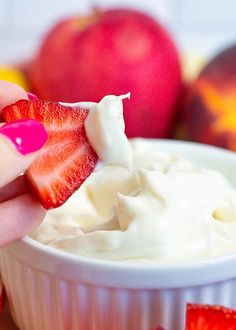 a hand holding a strawberry over a bowl of whipped cream with apples in the background