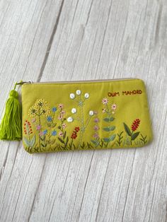 a yellow embroidered pencil case with flowers and tassels on the front, sitting on a white wooden surface