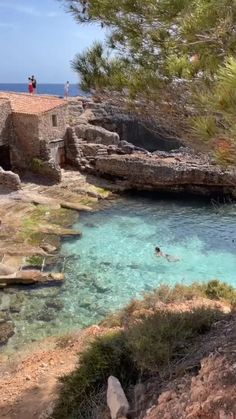 people are swimming in the clear blue water near some rocks and trees, while one person is standing on top of an old building