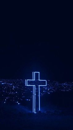 a cross lit up in the dark on top of a hill with city lights behind it