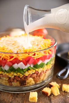 a person pouring dressing into a glass bowl filled with vegetables and other foods on top of a wooden table