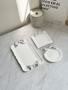 three white plates and two silver trays sitting on a marble counter top next to a candle