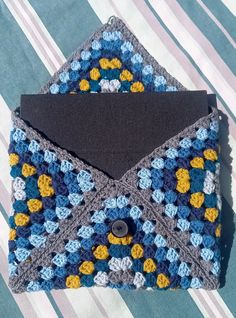 a blue and yellow crocheted envelope with a black button on the front, sitting on a striped cloth