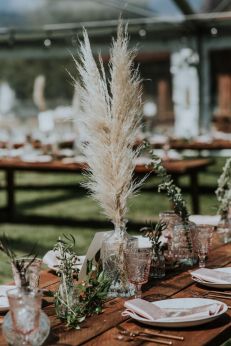 the table is set with white plates and silverware, tall pamodia grass in vases