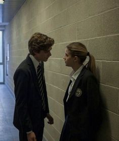 two people standing next to each other in front of a brick wall and looking at each other