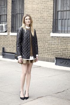 a woman standing in front of a brick building wearing a black jacket and tan skirt