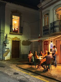 group of people sitting around a table in front of a building at night with lights on