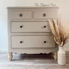 a white dresser sitting on top of a wooden floor next to a vase with dry grass in it