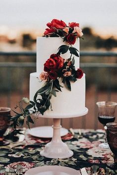 a white wedding cake with red flowers on top and greenery around the edges, sitting on a table outdoors