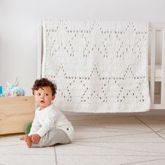 a baby sitting on the floor in front of a crib with a white blanket