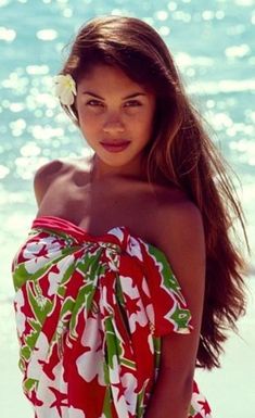 a beautiful young woman standing on top of a beach next to the ocean wearing a flower in her hair